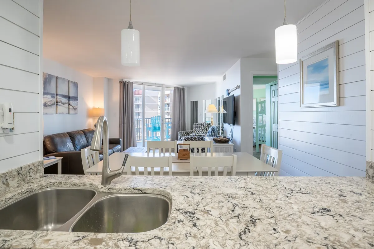 A bright open-concept space featuring a granite countertop, dining table, and a living room seating area, with the balcony and ocean visible in the background. The room is styled in a cheerful, contemporary manner that invites group activities and shared experiences.