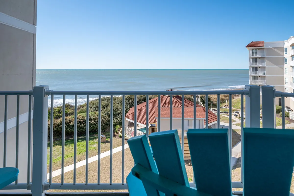 The image showcases a private balcony with cozy seating overlooking a sweeping ocean view. Below, lush landscaped gardens stretch out, creating a tranquil oasis between the condo and the beach. The soft morning light and serene waves evoke a peaceful, relaxing atmosphere, perfect for unwinding or enjoying coffee as the sun rises.