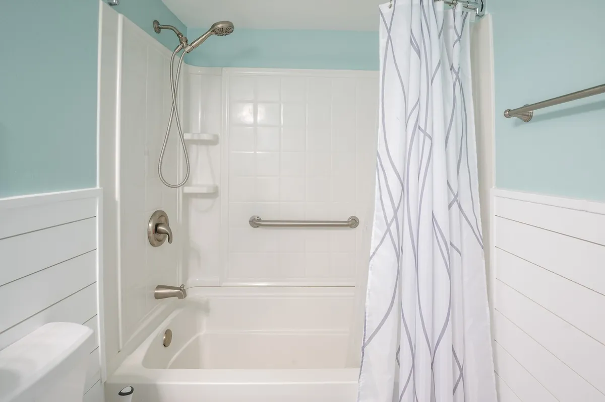 A bright, calming bathroom with a bathtub, showerhead, white paneling, and a shower curtain. The space is designed for relaxation and practical routines, evoking a sense of refreshment and tranquility.