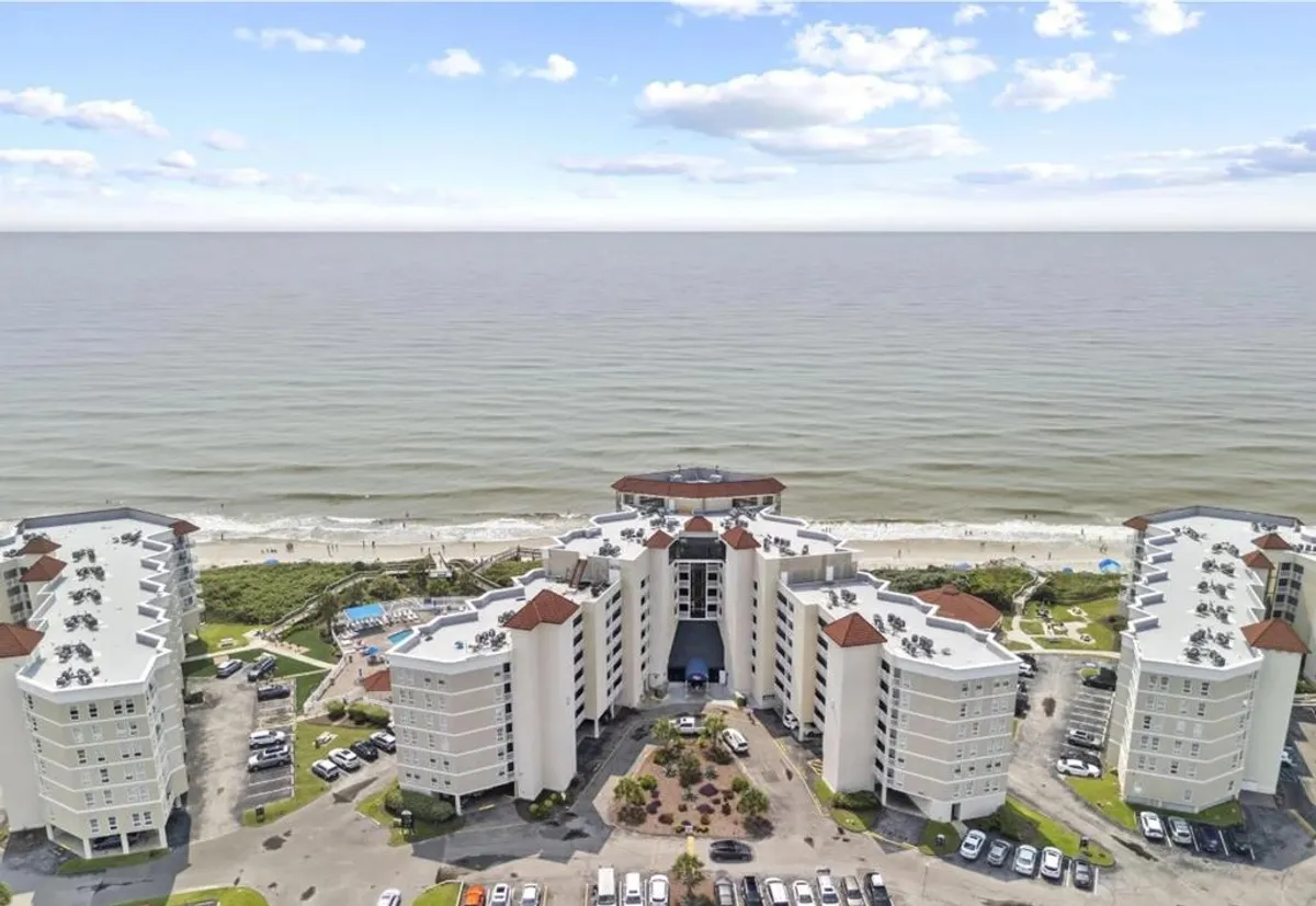 A wide exterior view showing the oceanfront pool area, landscaped gardens, and the buildings beyond, all overlooking the ocean. Guests are visible enjoying the pool, sunbathing, and the scenic vistas, with easy access to the beach.