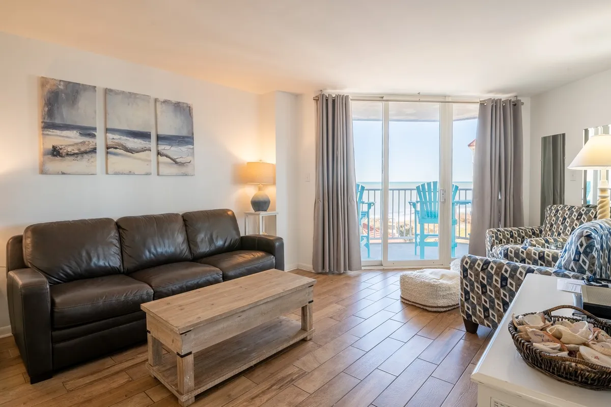 A cozy interior living area with a leather sofa, wooden coffee table, and a large window leading out to a balcony. The view through the window hints at a beautiful sunset or sunrise, blending interior comfort with outdoor scenery.