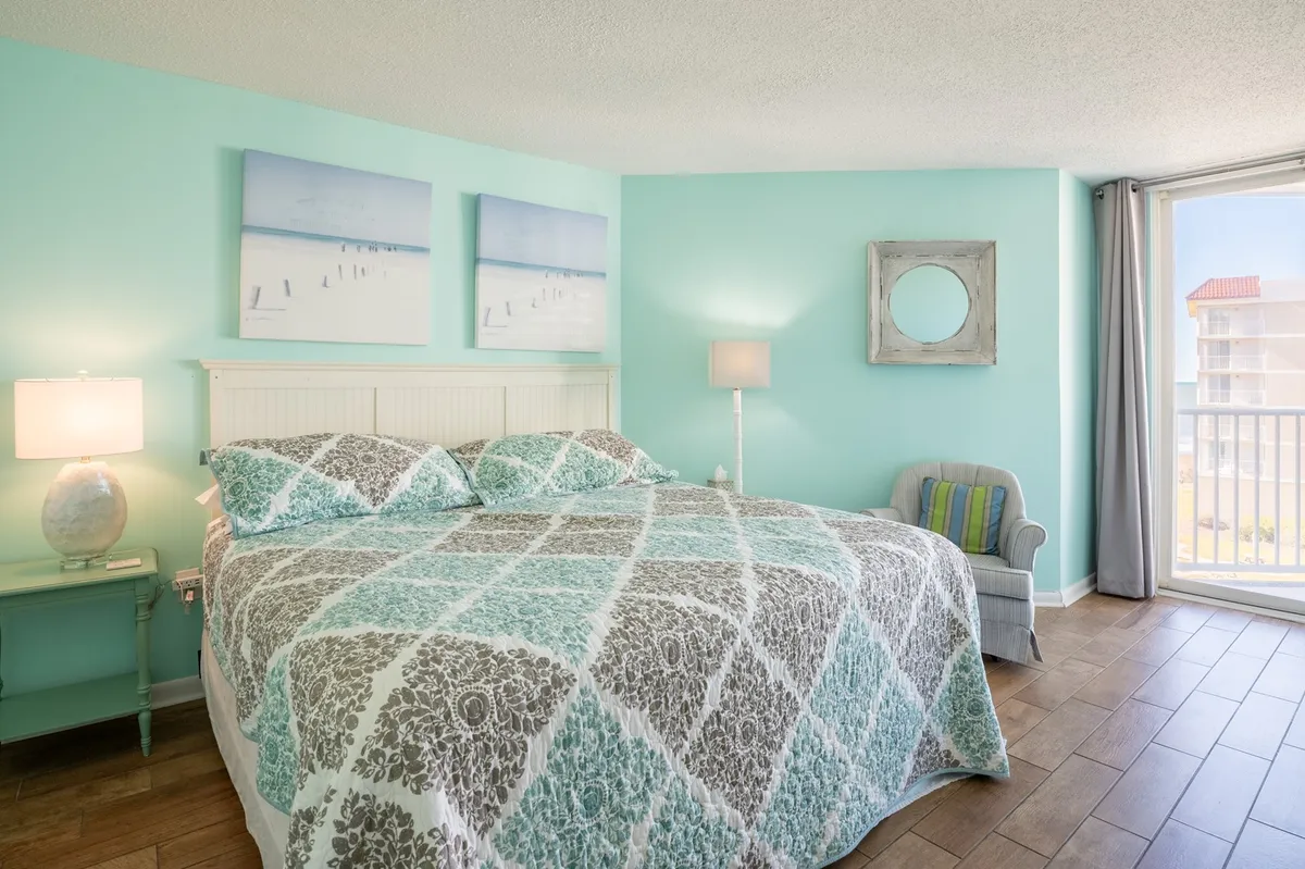 A serene bedroom with a patterned comforter on a king-sized bed, two beach-themed wall art pieces, and a large window allowing natural light to fill the space. The setting is calm, featuring soft finishes and a welcoming, restful ambiance.