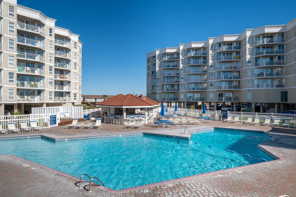 A sparkling outdoor swimming pool surrounded by lounge chairs and a poolside cabana, with buildings in the background. The area is set up for sunbathing, swimming, and socializing, reflecting a lively and inviting poolside atmosphere.
