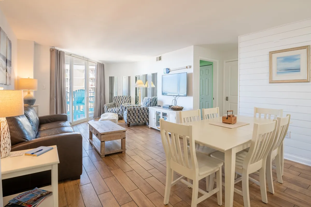 A bright, welcoming living and dining area featuring a comfortable leather sofa, white dining chairs, colorful accent chairs, and large windows that flood the space with natural light. The setup is ideal for casual meals, socializing, or relaxing after a day at the beach.