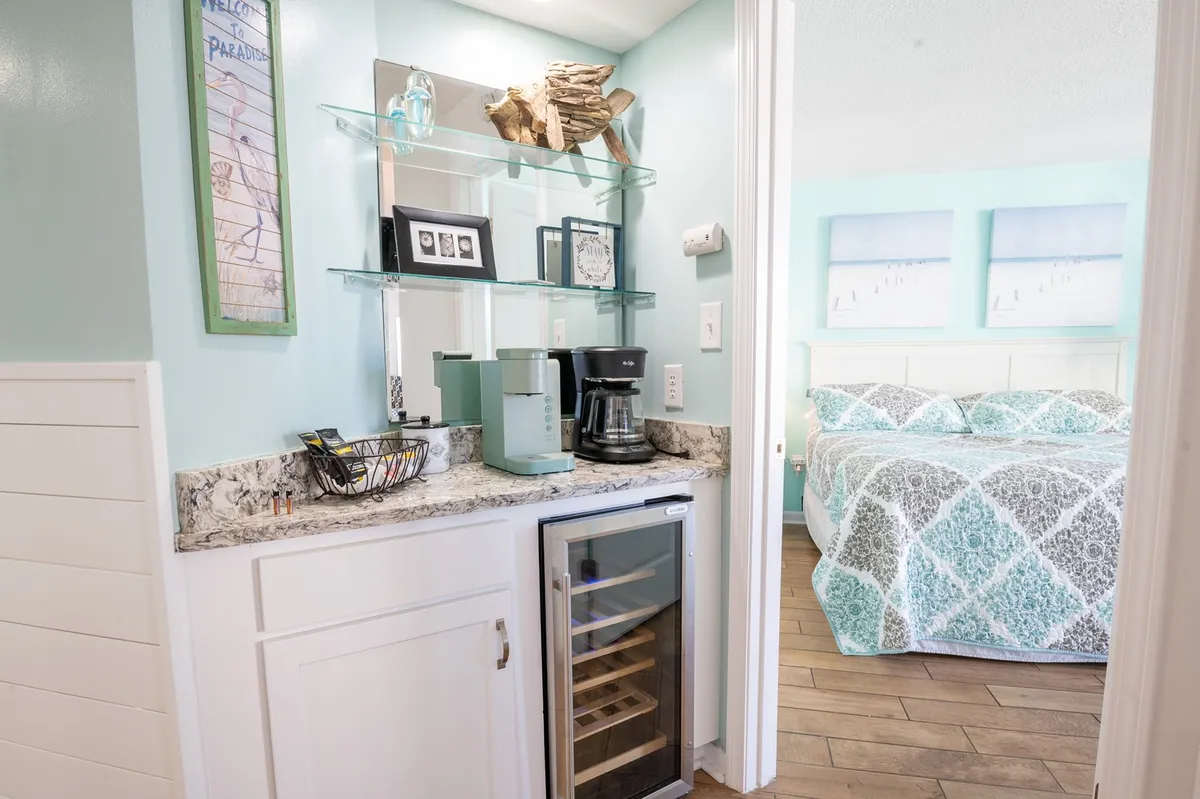 This image captures a cozy interior scene featuring a coffee maker and wine fridge set against a backdrop of decorative driftwood and a bed with patterned bedding. The space invites guests to prepare their morning coffee while enjoying views of the sea, suggesting both self-catering convenience and a comfortable, home-like atmosphere.